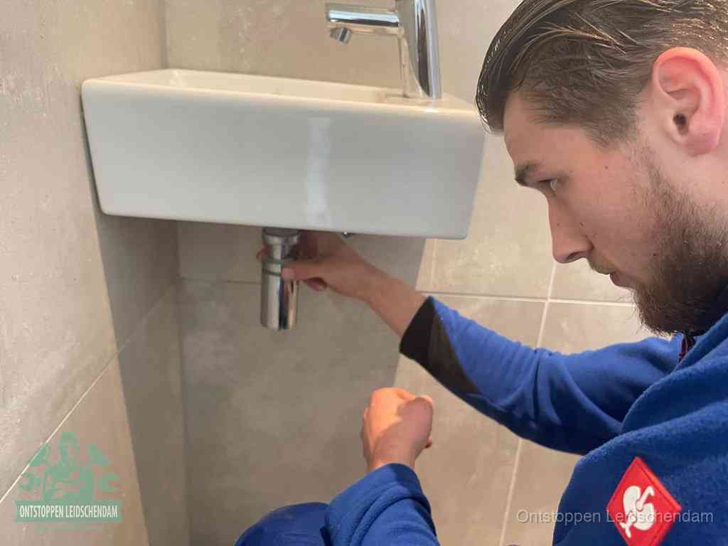 Een monteur draait de sifon onder een fonteinwasbak los om de wasbak te ontstoppen in Leidschendam